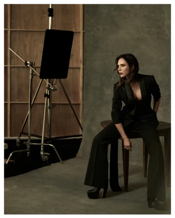 Candid studio portrait of a stylish adult woman in a black suit, seated on a stool, with studio lighting and neutral backdrop.