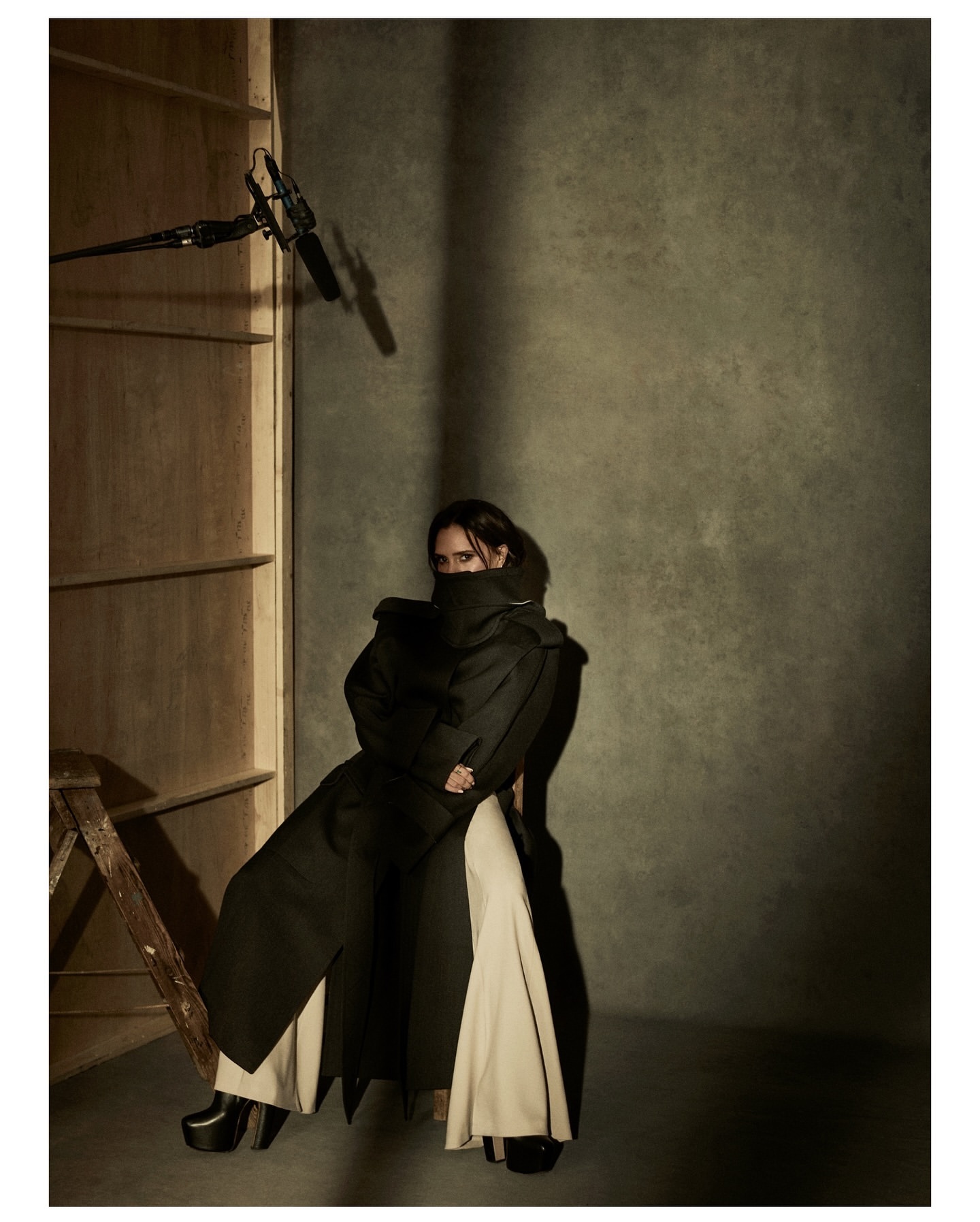 A person posing in an overcoat and wide-legged pants in a studio setting with a boom mic and ladder visible, with a neutral color palette.
