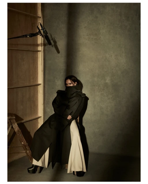 A person posing in an overcoat and wide-legged pants in a studio setting with a boom mic and ladder visible, with a neutral color palette.