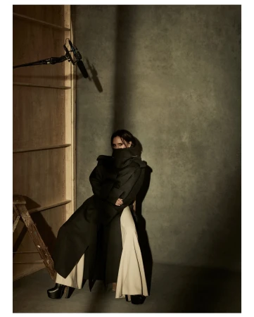 A person posing in an overcoat and wide-legged pants in a studio setting with a boom mic and ladder visible, with a neutral color palette.