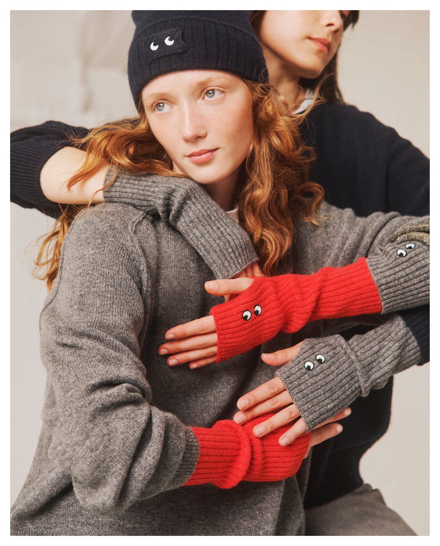 Two models in a neutral studio setting, one with red fingerless gloves with eyes, the other with a knit cap with eyes.