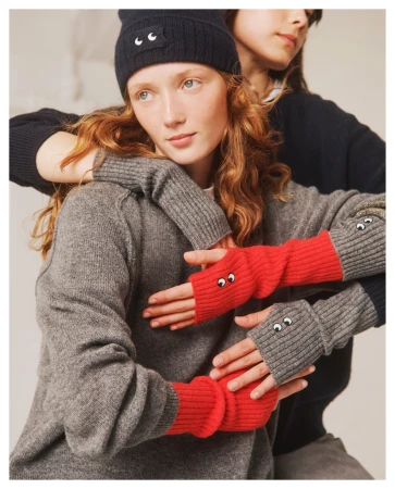 Two models in a neutral studio setting, one with red fingerless gloves with eyes, the other with a knit cap with eyes.