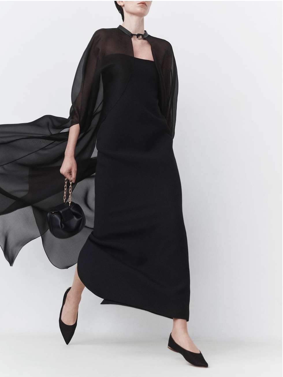 Elegant woman in a black dress with a sheer cape and matching flats carries a small black purse against a white backdrop.