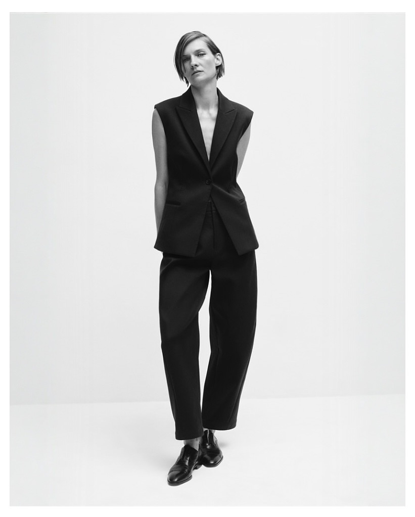 Black and white fashion shot of a stylish adult woman posing in a sleeveless black suit with dress pants and shiny black shoes.