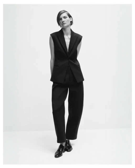 Black and white fashion shot of a stylish adult woman posing in a sleeveless black suit with dress pants and shiny black shoes.