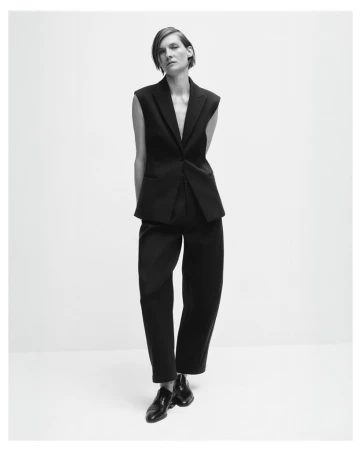Black and white fashion shot of a stylish adult woman posing in a sleeveless black suit with dress pants and shiny black shoes.