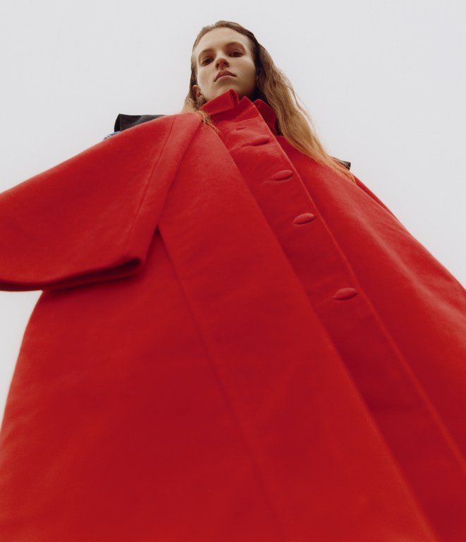 Low-angle shot of a fair-skinned adult woman in a red collared coat against a bright white backdrop, fashion and editorial style.