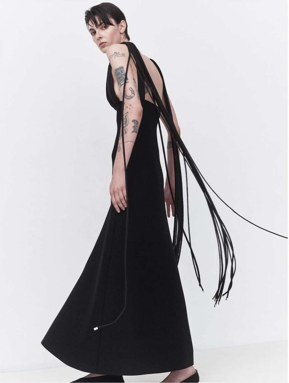 Elegant woman in a long black dress with flowing black cords poses in a white studio. Tattoos are visible on her left arm.