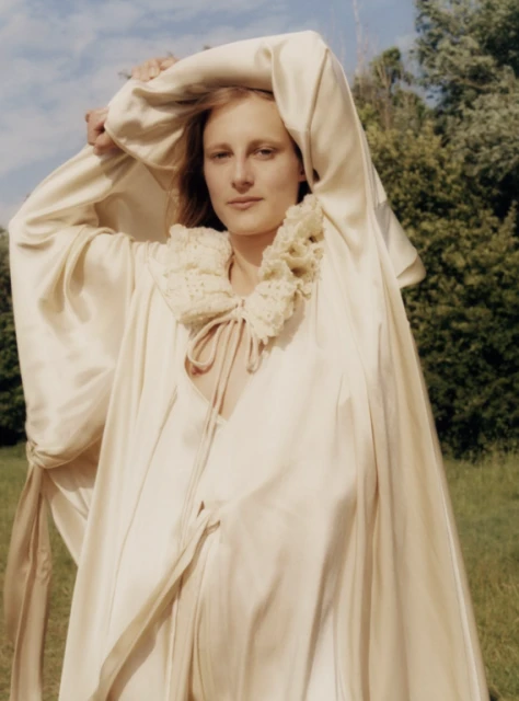 Portrait of an adult woman wearing a cream-colored robe with a ruffled collar, posed outdoors in a bright, soft natural light.