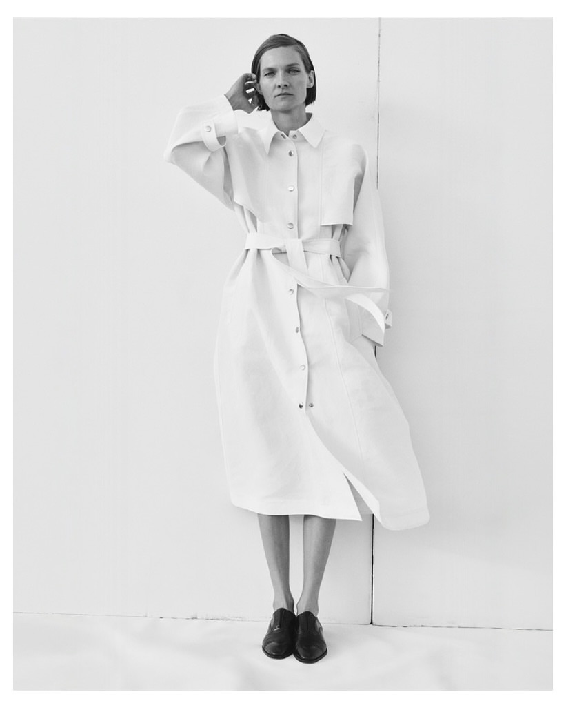 A woman in a belted dress with black shoes stands against a white wall in a minimalist studio setting, casting a subtle shadow.