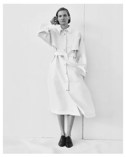 A woman in a belted dress with black shoes stands against a white wall in a minimalist studio setting, casting a subtle shadow.