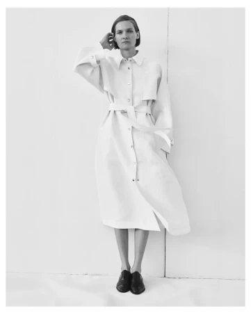 A woman in a belted dress with black shoes stands against a white wall in a minimalist studio setting, casting a subtle shadow.