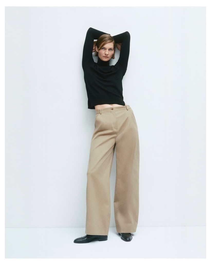 Woman in a black turtleneck and wide-leg pants poses in front of a white wall in a studio setting, showcasing a modern fashion look.