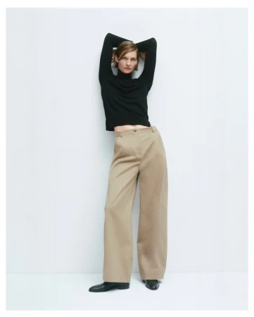 Woman in a black turtleneck and wide-leg pants poses in front of a white wall in a studio setting, showcasing a modern fashion look.