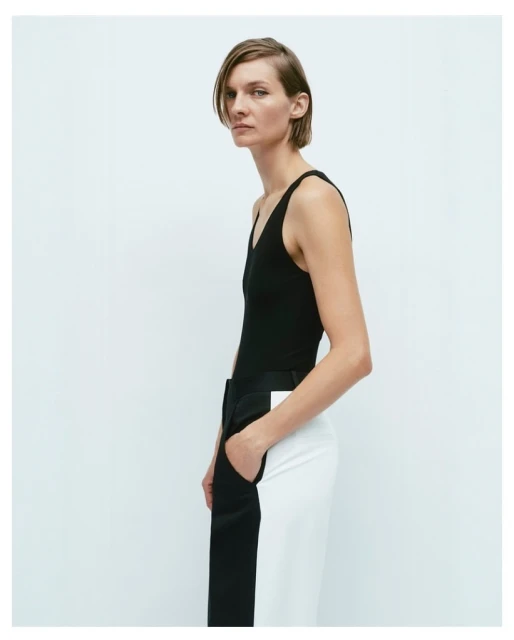 A woman stands in front of a white background wearing a black sleeveless top and color-blocked black-and-white pants.