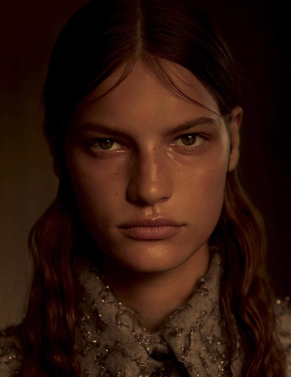 Close-up portrait of a woman with long brown hair wearing an embellished grey collared shirt against a dark moody background.