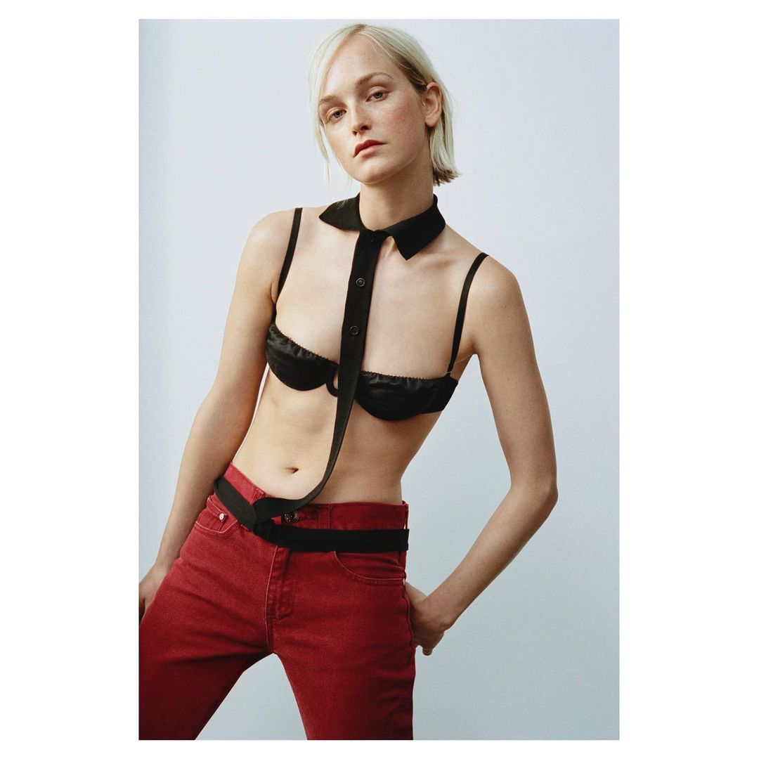 Young woman in fashion-forward red jeans and a black bra top with a unique collar posing in a brightly lit, neutral-toned studio setting.