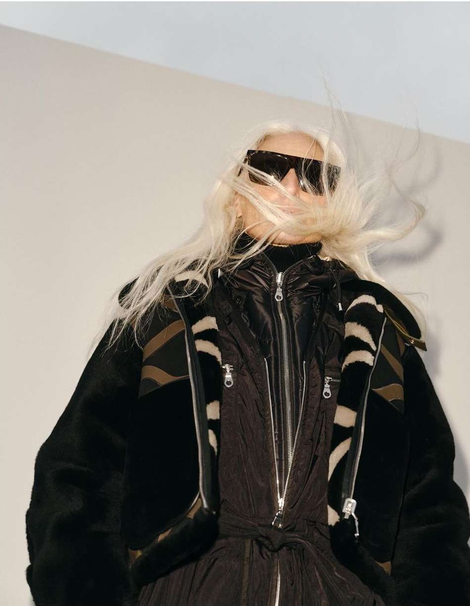 A fair-skinned woman with long blond hair and large sunglasses, wearing a black jacket, poses in front of a light-colored background.