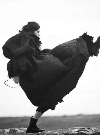 Black and white editorial fashion image of a model wearing a voluminous quilted coat and dark boots kicking high outdoors.