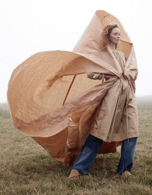Woman in a tan paper garment and blue jeans stands in a grassy field on a misty day, posing with an avant-garde sculptural silhouette.