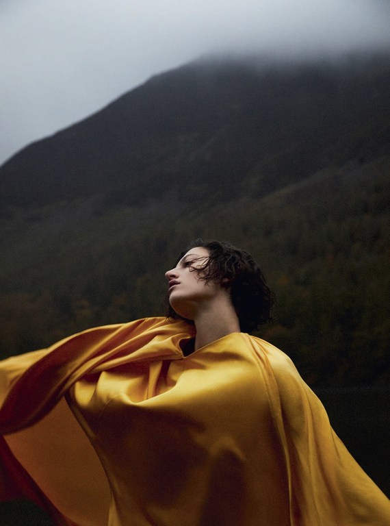 Woman in flowing golden robe poses outdoors with a mountain background against a cloudy sky, creating a dramatic, artistic scene.