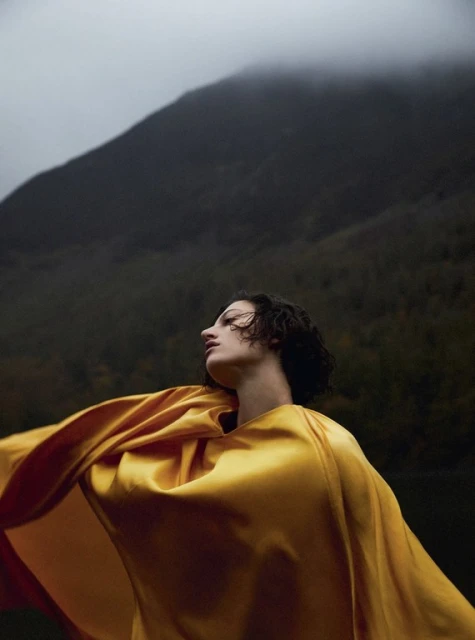 Woman in flowing golden robe poses outdoors with a mountain background against a cloudy sky, creating a dramatic, artistic scene.