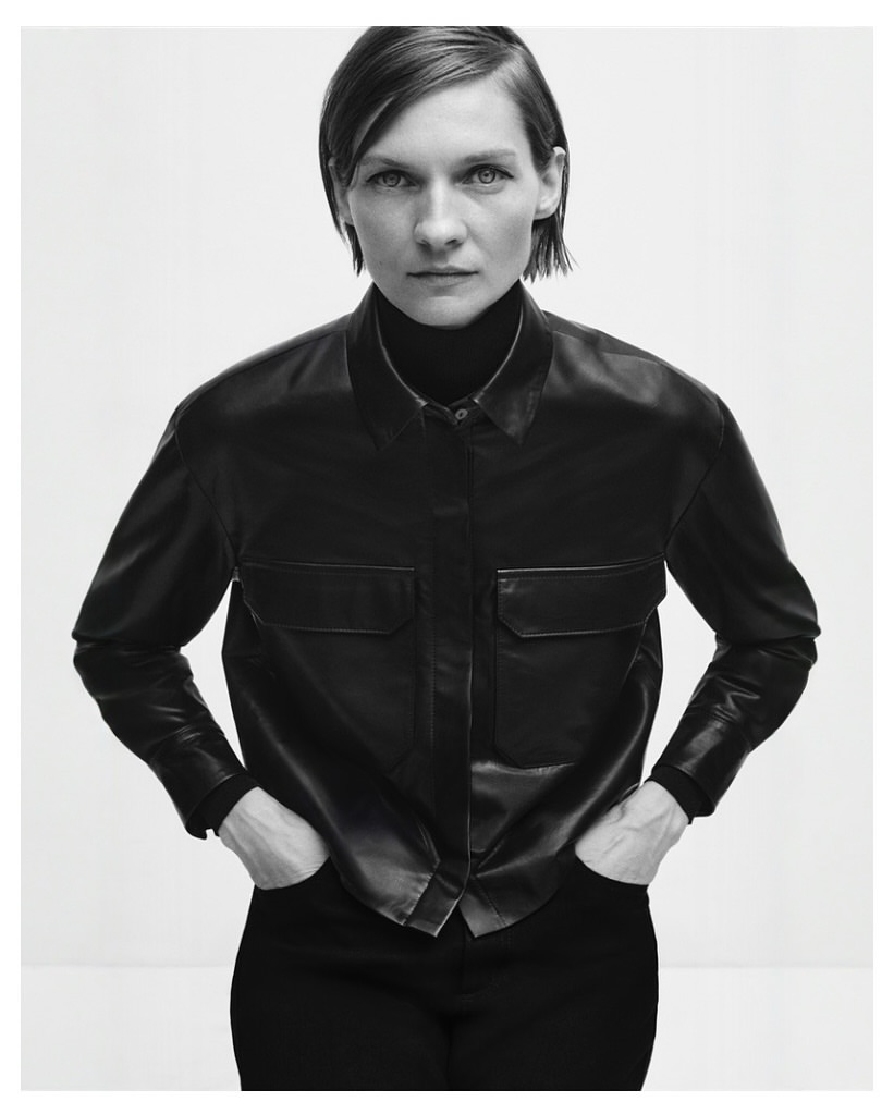 Monochrome studio shot of a woman in a black button-down shirt with hands in pockets, standing against a white background.