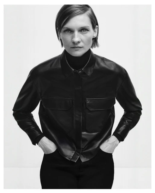 Monochrome studio shot of a woman in a black button-down shirt with hands in pockets, standing against a white background.