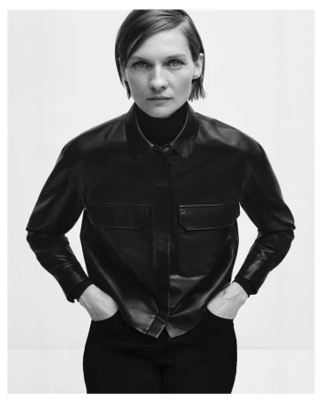 Monochrome studio shot of a woman in a black button-down shirt with hands in pockets, standing against a white background.