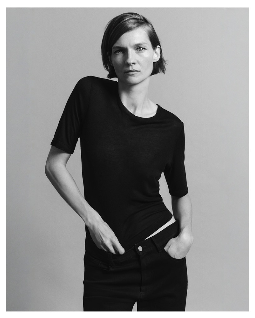 Model with short dark hair wearing a black short-sleeved top against a plain grey studio backdrop