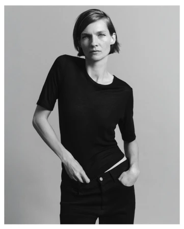 Model with short dark hair wearing a black short-sleeved top against a plain grey studio backdrop