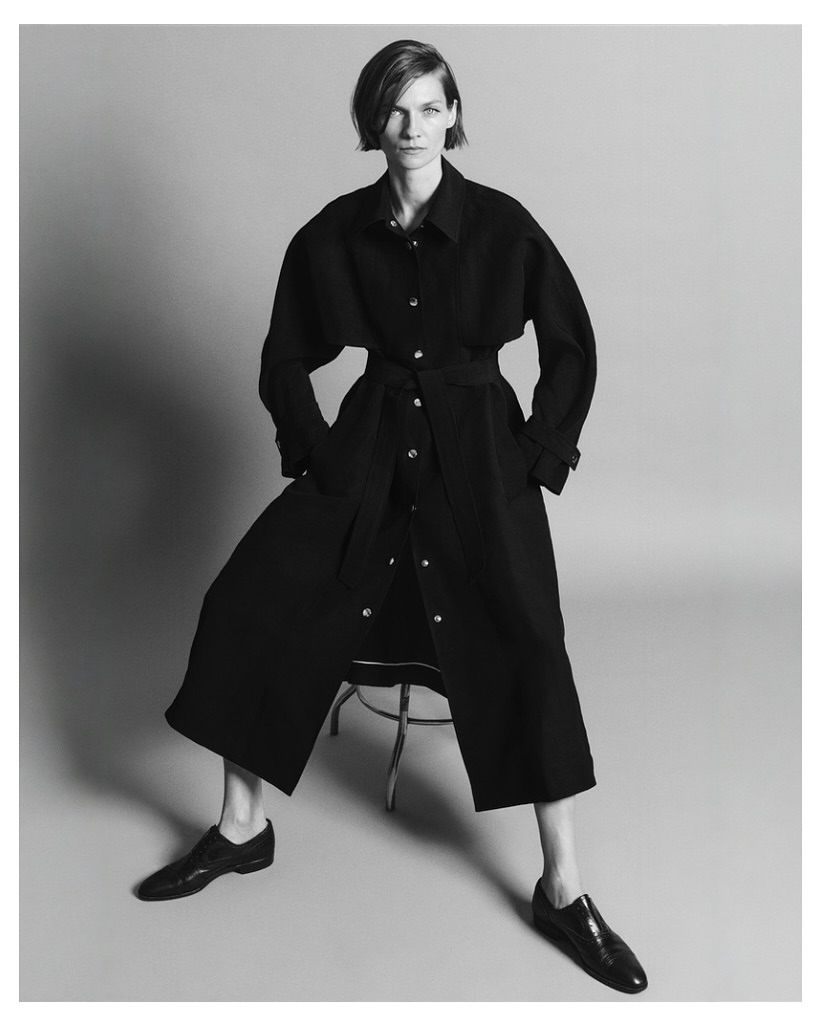 Monochrome studio shot of a stylish adult female model in a belted black coat dress, seated on a stool with black leather shoes.