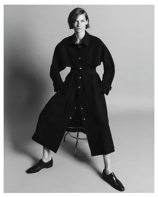Monochrome studio shot of a stylish adult female model in a belted black coat dress, seated on a stool with black leather shoes.