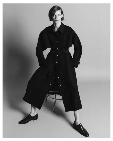 Monochrome studio shot of a stylish adult female model in a belted black coat dress, seated on a stool with black leather shoes.
