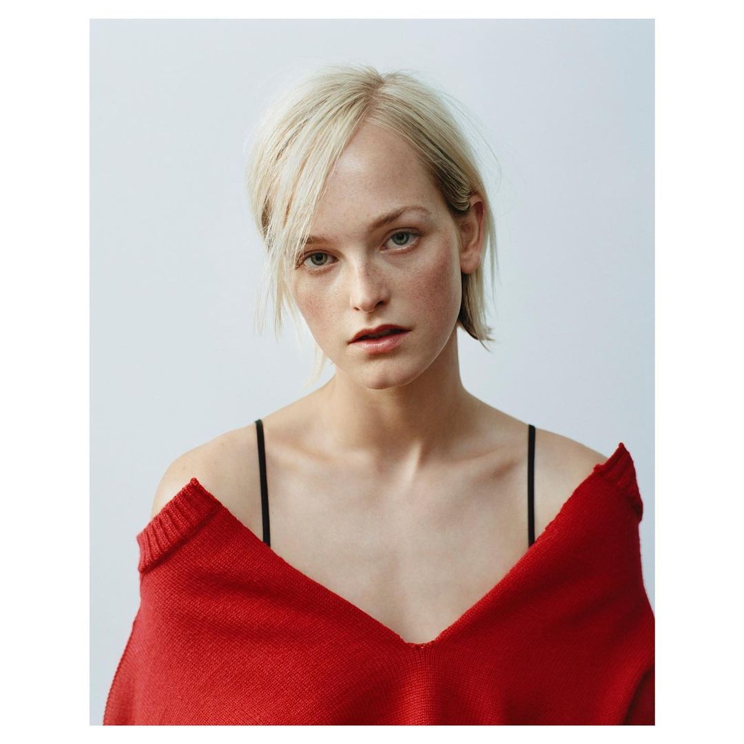 Portrait of a blonde woman wearing an off-the-shoulder red knit sweater against a plain white background.
