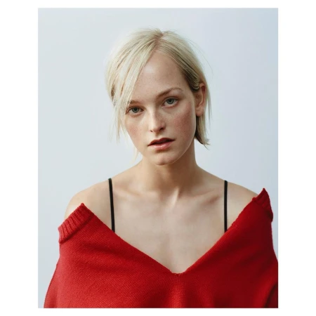 Portrait of a blonde woman wearing an off-the-shoulder red knit sweater against a plain white background.