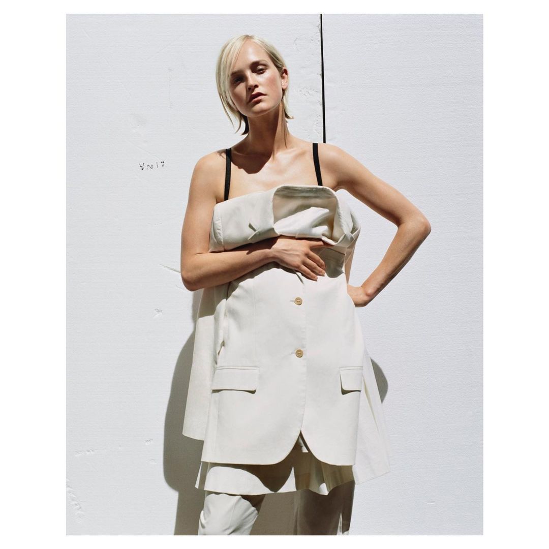 A woman stands with her hand on her hip wearing a cream-colored suit dress in front of a white wall, posing in the daytime in the studio.