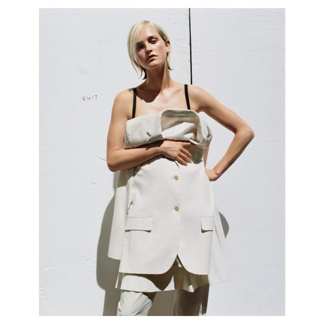A woman stands with her hand on her hip wearing a cream-colored suit dress in front of a white wall, posing in the daytime in the studio.