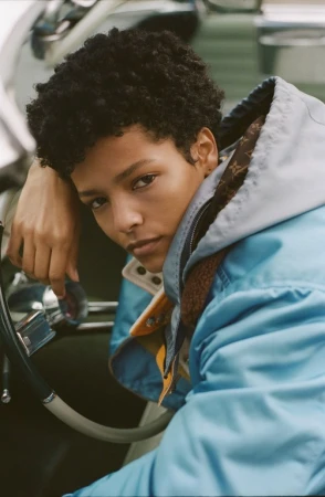 Close-up portrait of a model with short dark curly hair wearing a blue jacket leaning against a vintage car interior