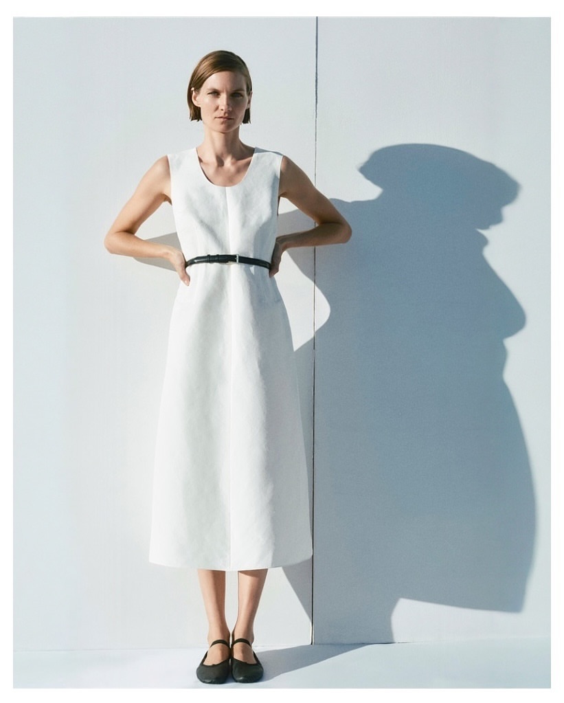 An adult woman stands with hands on her hips, wearing a sleeveless white dress with a black belt, against a white wall in bright light.
