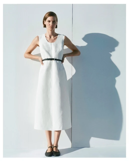 An adult woman stands with hands on her hips, wearing a sleeveless white dress with a black belt, against a white wall in bright light.