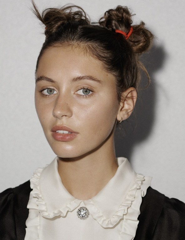 Portrait of a young woman with fair skin, light eyes, and two bun hairstyle wearing a white ruffled collared shirt against a plain backdrop.