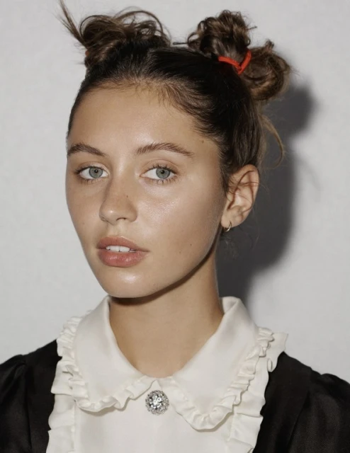 Portrait of a young woman with fair skin, light eyes, and two bun hairstyle wearing a white ruffled collared shirt against a plain backdrop.
