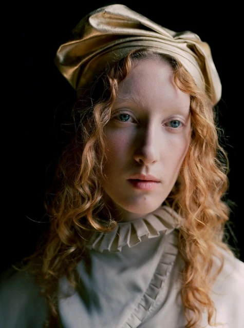 Portrait of a pale-skinned woman with curly strawberry blonde hair wearing a gold headwrap and a high-necked ivory dress in a studio.