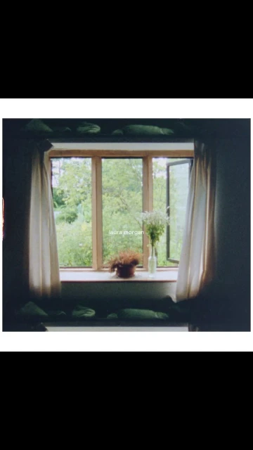 Indoor view of an open window with curtains and flowers, overlooking a lush green outdoor scene, creating a tranquil and inviting atmosphere.