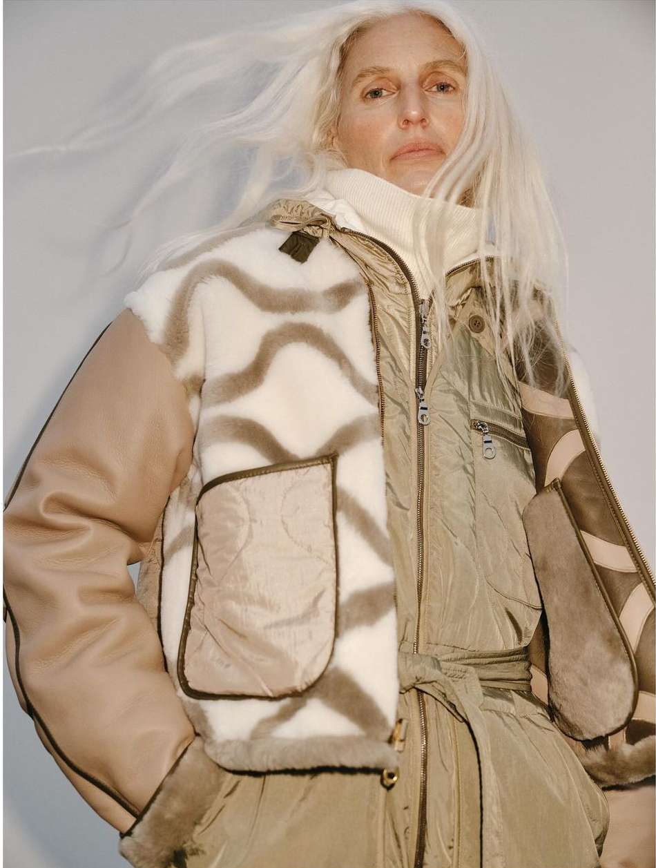 Mature woman poses in a studio wearing a light-brown jacket with a mixed pattern and texture. She has white hair and fair skin.