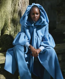 Lupita Nyong'o seated outdoors wearing a bright blue hooded coat with voluminous sleeves, styled by Cathy Kasterine.