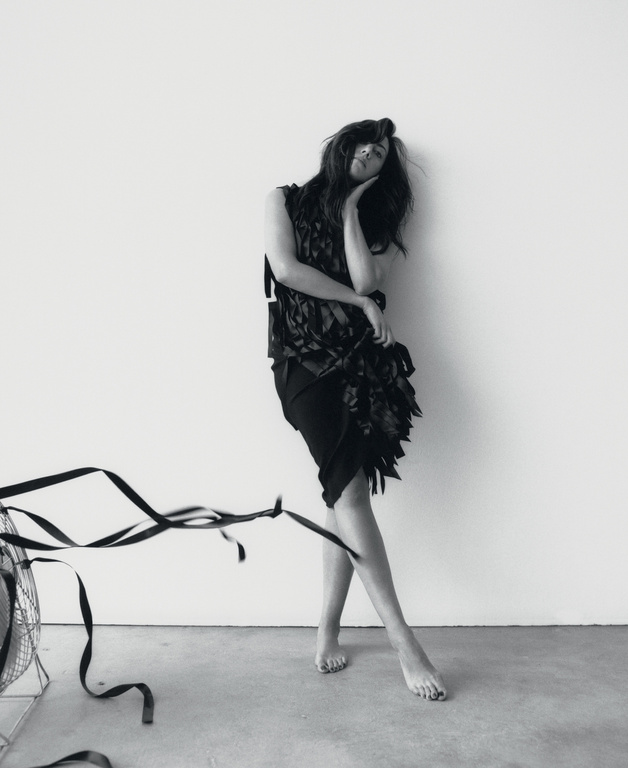 Black-and-white image of a woman in a dark dress leaning against a wall, with a vintage fan and ribbon visible in the foreground.