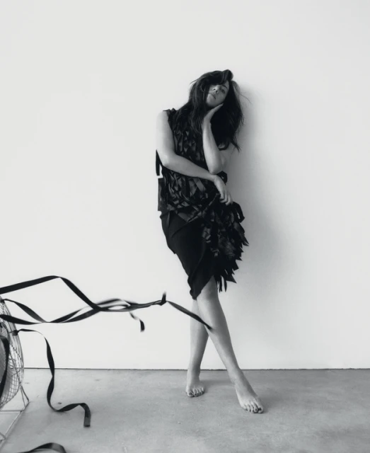 Black-and-white image of a woman in a dark dress leaning against a wall, with a vintage fan and ribbon visible in the foreground.