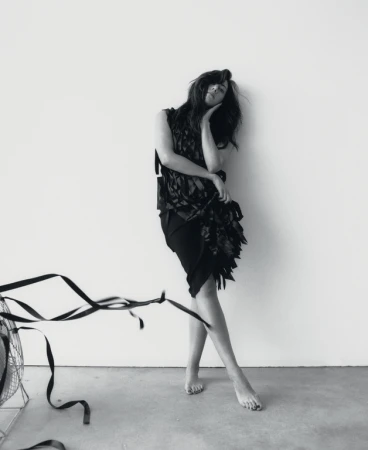 Black-and-white image of a woman in a dark dress leaning against a wall, with a vintage fan and ribbon visible in the foreground.